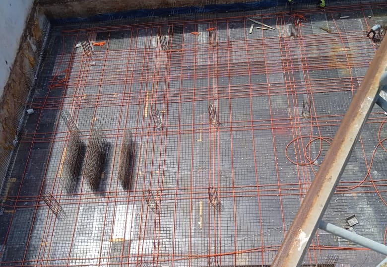 A detailed aerial close-up of a construction site showing steel reinforcement mesh covered with orange unbonded post-tensioning cables (tendons) laid out on formwork for a post-tension slab.